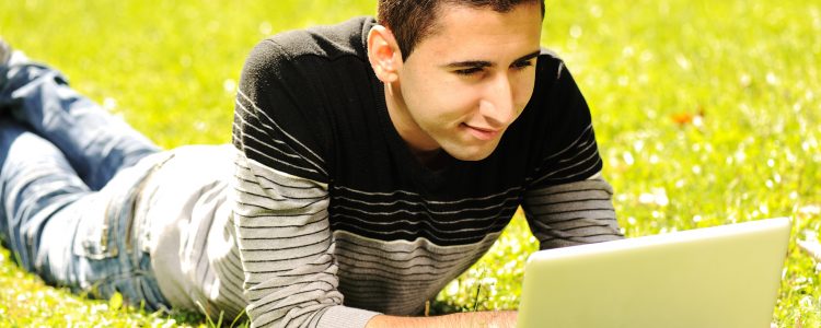 Happy young man laying on green grass in nature and working on laptop
