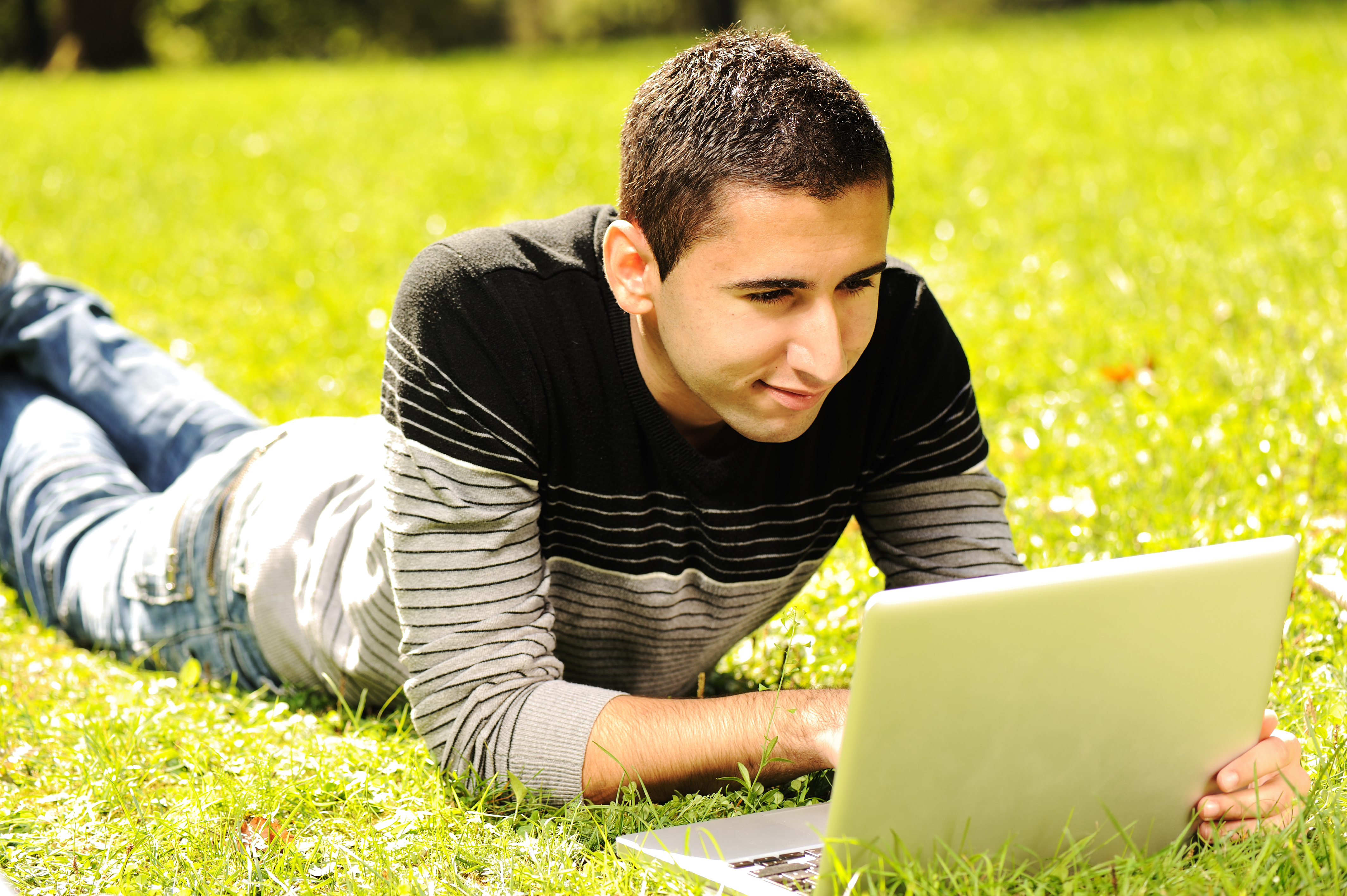 Happy young man laying on green grass in nature and working on laptop